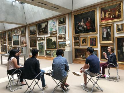 Museum docent and visitors sit in a semi-circle on stools in a gallery hung floor-to-ceiling with paintings.