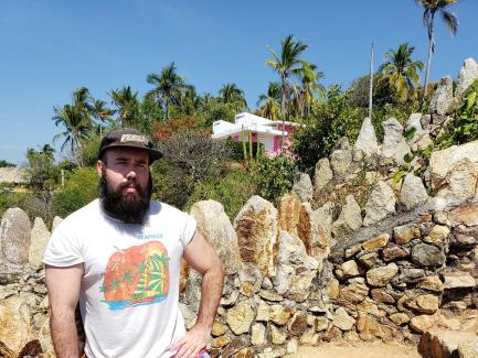 Man with a beard, wearing a white graphic t shirt andblack cap stands against a rocky wall with palm trees in background.