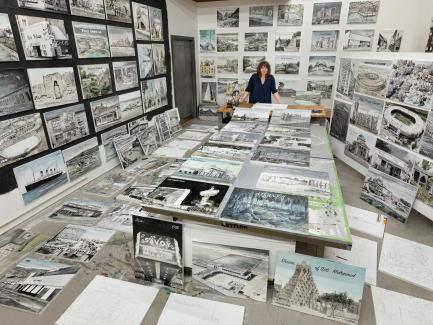 A white woman stands at a desk in a studio surrounded by artworks